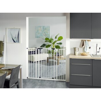 Ein weißes Haustiergitter steht in einer offenen Tür, die eine moderne Küche von einem Wohnbereich mit einer grünen Pflanze und einem hellen Sofa trennt.