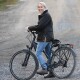 Person in schwarzer Jacke und blauen Jeans steht mit einem schwarzen Fahrrad auf einem Kiesweg im Freien.