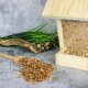 Ein hölzerner Vogelhäuschen, gefüllt mit Samen, mit einer Schaufel Samen, die daneben verschüttet ist, vor einem strukturierten grauen Hintergrund und grünem Gras.
