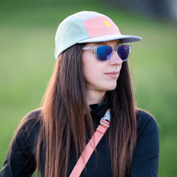Person mit verschwommenem Gesicht, trägt pastellfarbene Kappe und Sonnenbrille, lange braune Haare, schwarze Jacke und rosa Schulterriemen