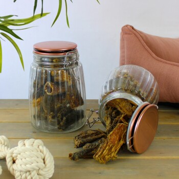 Zwei Glasbehälter gefüllt mit Haustierleckereien, einer umgekippt mit herausfallenden Leckereien, auf einem Holztisch neben einem Seilspielzeug und einem rosa Kissen.