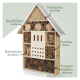 Ein hölzernes Insektenhotel in Form eines Hauses, gefüllt mit Holzspänen, Bambus und Tannenzapfen, beschriftet auf Deutsch.