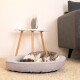 Getigerte Katze schläft eingerollt in einem runden, grauen, plüschigen Haustierbett auf beigem Teppich neben einem Beistelltisch im Mid-Century-Stil und einem grauen Sofa.