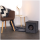 Gemütlicher Wohnraum mit einem braunen Stuhl, gemustertem Kissen, runder Tisch, Lampe und einer grauen Katze, die aus einem schwarzen Filzquadrat schaut.