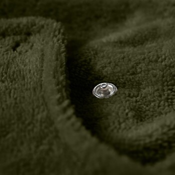 Ein brauner Hund, der auf einem sandigen Strand steht, trägt einen gemütlichen, übergroßen olivgrünen Hoodie, mit hohem Gras und einem Pfosten im Hintergrund.