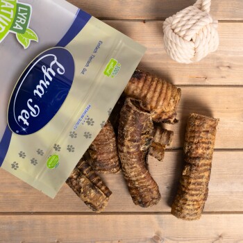 Eine Tüte Lyra Pet natürliche Hundeleckerlis mit mehreren gerollten, fleischigen Kaustangen, die auf einer Holzoberfläche präsentiert werden, mit einem Seilspielzeug in der Nähe.