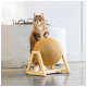 Un chat marron duveteux se tient sur un grattoir en bois avec une balle ronde recouverte de corde, dans un intérieur minimaliste.
