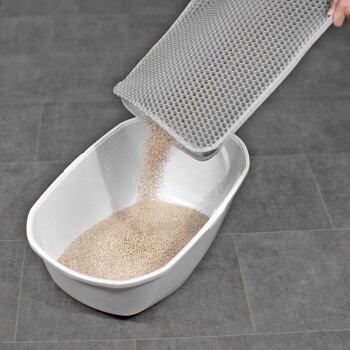 Eine Person, die Katzenstreu von einer grauen Schaufel in eine weiße Katzentoilette auf einem grauen Fliesenboden schüttet.