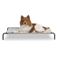 Ein flauschiger brauner und weißer Hund, der auf einem grauen erhöhten Haustierbett mit schwarzen Metallbeinen entspannt, vor einem weißen Hintergrund.