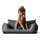 Ein brauner Hund sitzt aufrecht auf einem plüschigen grauen Haustierbett mit zwei weichen Kissen und schaut neugierig zur Seite.