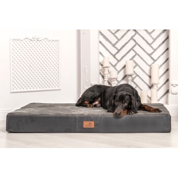 Ein schwarz-brauner Hund ruht auf einem plüschigen grauen Haustierbett mit einem Lederanhänger, der mit "tierland" beschriftet ist. Der Hintergrund zeigt weiße Kerzen und eine dekorative Wand.