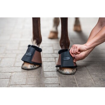Eine Person befestigt einen schwarzen und braunen Hufschuh mit einem flauschigen Innenfutter am Vorderhuf eines Pferdes, der das Design des Produkts zeigt.