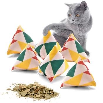 Eine graue Katze interagiert verspielt mit bunten dreieckigen Katzenminze-Kissen, mit getrockneter Katzenminze, die auf der weißen Oberfläche verstreut ist.