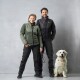 Ein Mann und eine Frau stehen nebeneinander, beide tragen stilvolle Outdoor-Jacken und Hosen, mit einem Golden Retriever, der neben ihnen sitzt.