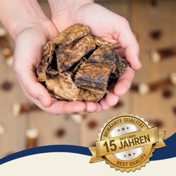 Ein Paar Hände, die große, getrocknete Stücke von Haustier-Kauartikeln halten, mit einem Abzeichen, das "Beste Qualität" und "15 Jahre" auf Deutsch angibt.