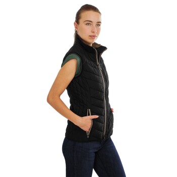 Eine Frau trägt eine schwarze ärmellose Steppweste über einem grünen Shirt und steht mit den Händen in den Taschen der Weste.