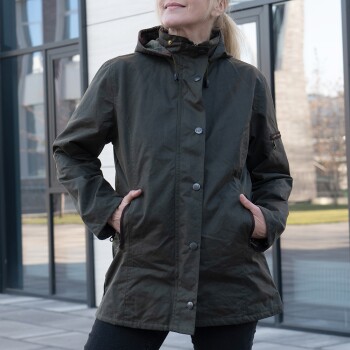 Frau trägt eine dunkelgrüne Kapuzenjacke mit Händen in den Taschen, steht draußen in der Nähe von Glasfenstern.