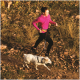 Eine Person in einer pinken Jacke joggt neben einem weißen Hund auf einem felsigen Pfad, umgeben von hohem Gras.