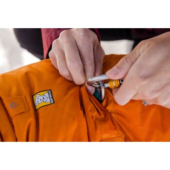 Eine Nahaufnahme einer Person, die einen Karabiner an einer orangefarbenen Hundejacke mit einem Ruffwear-Logo befestigt, und das sichere Design der Jacke zeigt.