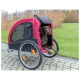 Ein brauner Hund sitzt bequem in einem roten und schwarzen Haustieranhänger mit Netzfenstern, der auf einem gepflasterten Weg in einem Grasbereich geparkt ist.