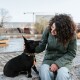 Lockighaarige Frau in grüner Jacke streichelt einen kleinen schwarzen Hund mit einem Halsband mit orangefarbener Quaste auf einer steinernen Uferkante.