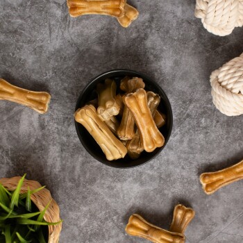 Eine schwarze Schüssel gefüllt mit verschiedenen hundeknochenförmigen Leckerlis in Brauntönen, umgeben von verstreuten Leckerlis und einem Seilspielzeug auf einer grauen Oberfläche.