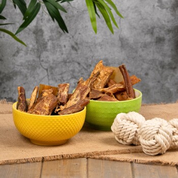 Zwei Glasbehälter mit Hundeleckerlis, einer umgekippt, mit einem geknoteten Seilspielzeug auf einem Holztisch und einem rosa Sofa im Hintergrund.