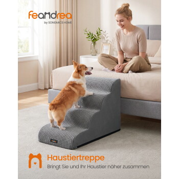 Corgi klettert graue Feandrea-Haustiertreppen zum Bett hinauf, während eine Frau lächelt; sichtbarer Text 'feandrea' und 'Haustiertreppe'.