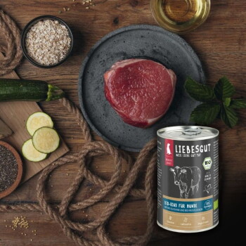 Dose LIEBESGUT-Hundefutter (Bio-Rind für Hunde) neben rohem Steak mit Zucchini, Haferflocken und Öl auf einem Holztisch.