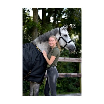 Eine Frau in einem grünen Shirt und schwarzen Jeans steht neben einem grauen Pferd, das eine schwarze Decke mit gelbem Rand trägt, in einer natürlichen Umgebung.