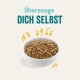 Miska z karmą dla zwierząt wypełniona małymi, okrągłymi granulkami, z towarzyszącym tekstem "Überzeuge DICH SELBST" w pogrubionych, kolorowych literach.