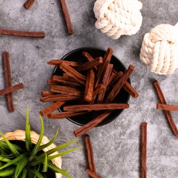 Eine schwarze Schüssel gefüllt mit braunen Haustierleckerlis, umgeben von verstreuten Leckerli-Sticks und zwei weißen Seilspielzeugen auf einer grauen Oberfläche.