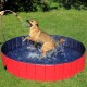 Ein glücklicher Hund spritzt in einem roten und blauen Haustierpool, während Wasser aus einem Schlauch gesprüht wird, den eine Person hält.
