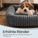 Schlafender Welpe in einem grauen, gerippten, runden Haustierbett mit cremefarbenem Knochenkissen; sichtbarer Text: 'Erhöhte Ränder'