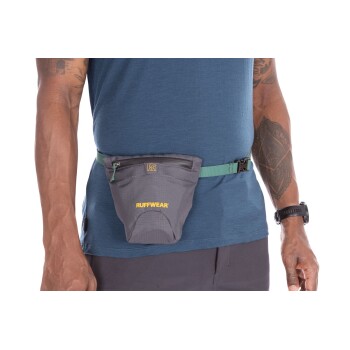 Eine Person in einem blauen Shirt und grauen Shorts wird mit einem grauen Ruffwear-Leckerli-Beutel gezeigt, der an einem grünen Gürtel um ihre Taille befestigt ist.