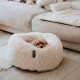 Ein kleiner Hund, der in einem flauschigen, runden Haustierbett auf einem hellen Teppich gekuschelt ist, mit einem gemütlichen Sofa im Hintergrund.