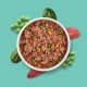Eine weiße Schüssel mit texturiertem braunem Tierfutter mit Brokkoli, umgeben von rohem Fleisch und Salat auf einem rosa Hintergrund.