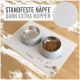 Graue Futternäpfe auf einer weißen Matte mit verstreutem Futter, mit dem Text "Standfeste Näpfe dank extra Noppen."