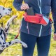 Eine Frau in einer marineblauen Jacke und schwarzen Leggings hält ein Smartphone, während ihr Dalmatiner hochreach. Sie trägt eine rote Bauchtasche vor herbstlichem Laub.