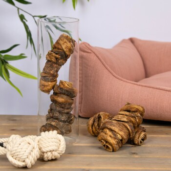 Eine Glasvase mit verdrehten Hundekauartikeln auf einem Holztisch, neben losen Kauartikeln, Seilspielzeugen und einem rosa Haustierbett im Hintergrund.