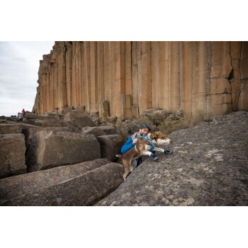 Eine Person hilft einem Hund, über ein felsiges Gelände in der Nähe von hohen, vertikalen Felsformationen zu klettern, mit anderen Wanderern im Hintergrund.