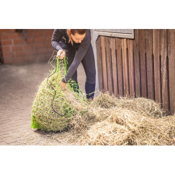 Eine Person in einem schwarzen Outfit hebt ein großes, grünes Heunetz, das mit Heu gefüllt ist, mit losem Heu, das auf dem Boden in einer Scheune verstreut ist.