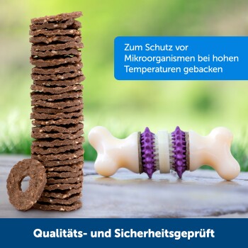 Braune Hundeleckerlis neben einem knochenförmigen Kauspielzeug, mit einer blauen Box, die "Für hohe Temperaturen gebacken" angibt, und einem Qualitätssicherungsbanner.