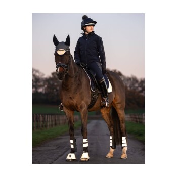 Ein Reiter in einer dunklen Jacke und einem Helm sitzt auf einem braunen Pferd, das weiße Beinschützer trägt, auf einem Landweg mit verschwommenem Hintergrund.