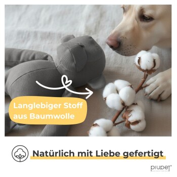 Hund schnüffelt an grauem Baumwollstoffspielzeug mit Baumwollpflanze; Text lautet "Langlebiger Stoff aus Baumwolle" und "Natürlich mit Liebe gefertigt."