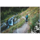 Ein Wanderer in tealfarbener Ausrüstung geht auf einem Pfad neben einem Wasserfall, begleitet von einem Hund mit einem Rucksack, umgeben von üppigem Grün.
