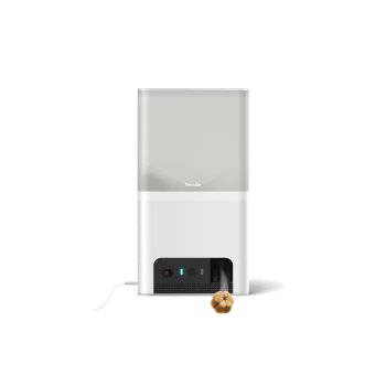 Eine moderne Petcube-Haustierkamera und Leckerli-Spender, mit einem eleganten weißen und grauen Design, aus dem ein Leckerli von unten ausgegeben wird.