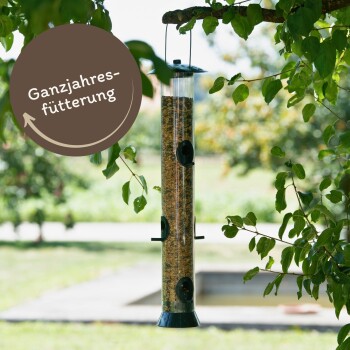 Ein Vogelhaus, das von einem Baum hängt, gefüllt mit Samen, mit einem runden Etikett, das 'Ganzjahresfütterung' (Jahresfütterung) liest.
