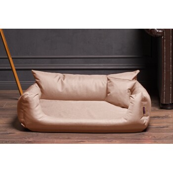 Ein plüschiges, beiges Haustierbett mit erhöhten Seiten und zwei weichen Kissen, vor einer dunklen Wand und einem Holzboden platziert.