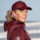 Eine Frau in einer kastanienbraunen Jacke und Mütze steht am Strand, mit Wellen im Hintergrund. Die Jacke hat ein Logo.